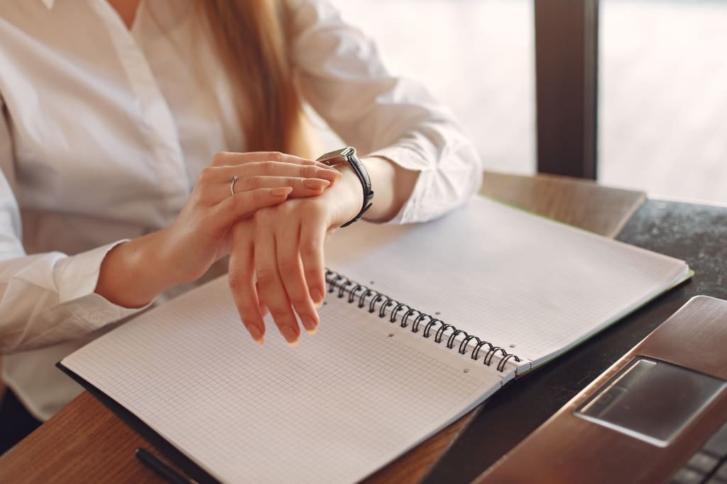 A person checking the time on their watch while sitting at a desk with an open notebook and a laptop beside them.